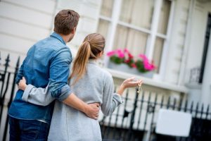 Couple with house key walking up to white home with window flower boxes.