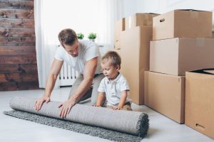 Father and son rolling up rug in front of boxes in preparation for move; relocating with chidlren after divorce has challenges.