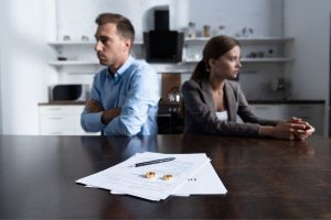 Couple sitting at table with backs to each other and forms, pen and wedding bands on table; common divorce mistakes